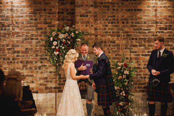 Bride puts grooms ring on his finger whilst celebrant looks on smiling